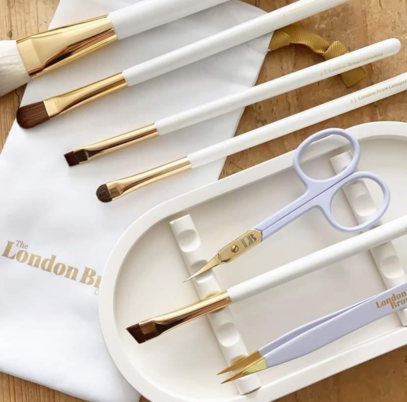 Set of white and gold makeup brushes with a pair of scissors on a wooden surface, branded 'The London Brow